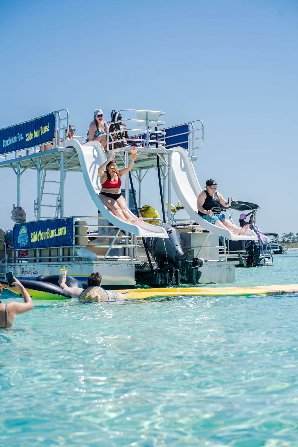 two people sliding off two waterslides on a double decker pontoon boat into the ocean